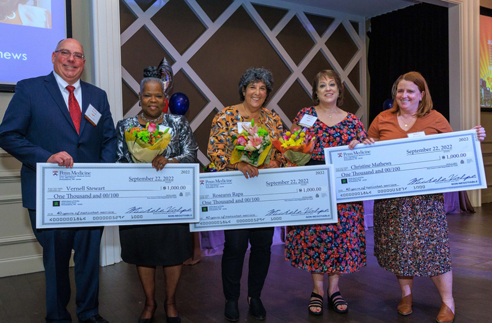 A man and four women stand in a row holding three oversized checks. Three of the women also hold bouquets of flowers.